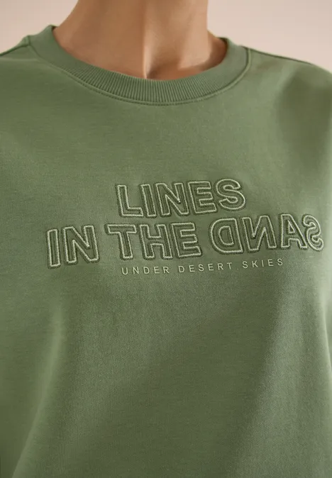 Olivgrünes T-Shirt mit dem aufgedruckten Schriftzug „LINES IN THE SAND“ und „UNDER DESERT SKIES“.